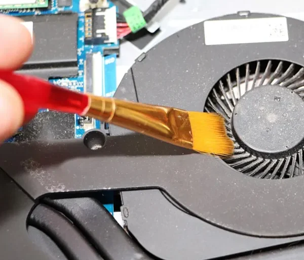 Technician cleaning a laptop cooling fan with a soft brush to remove internal dust as part of proper laptop maintenance.